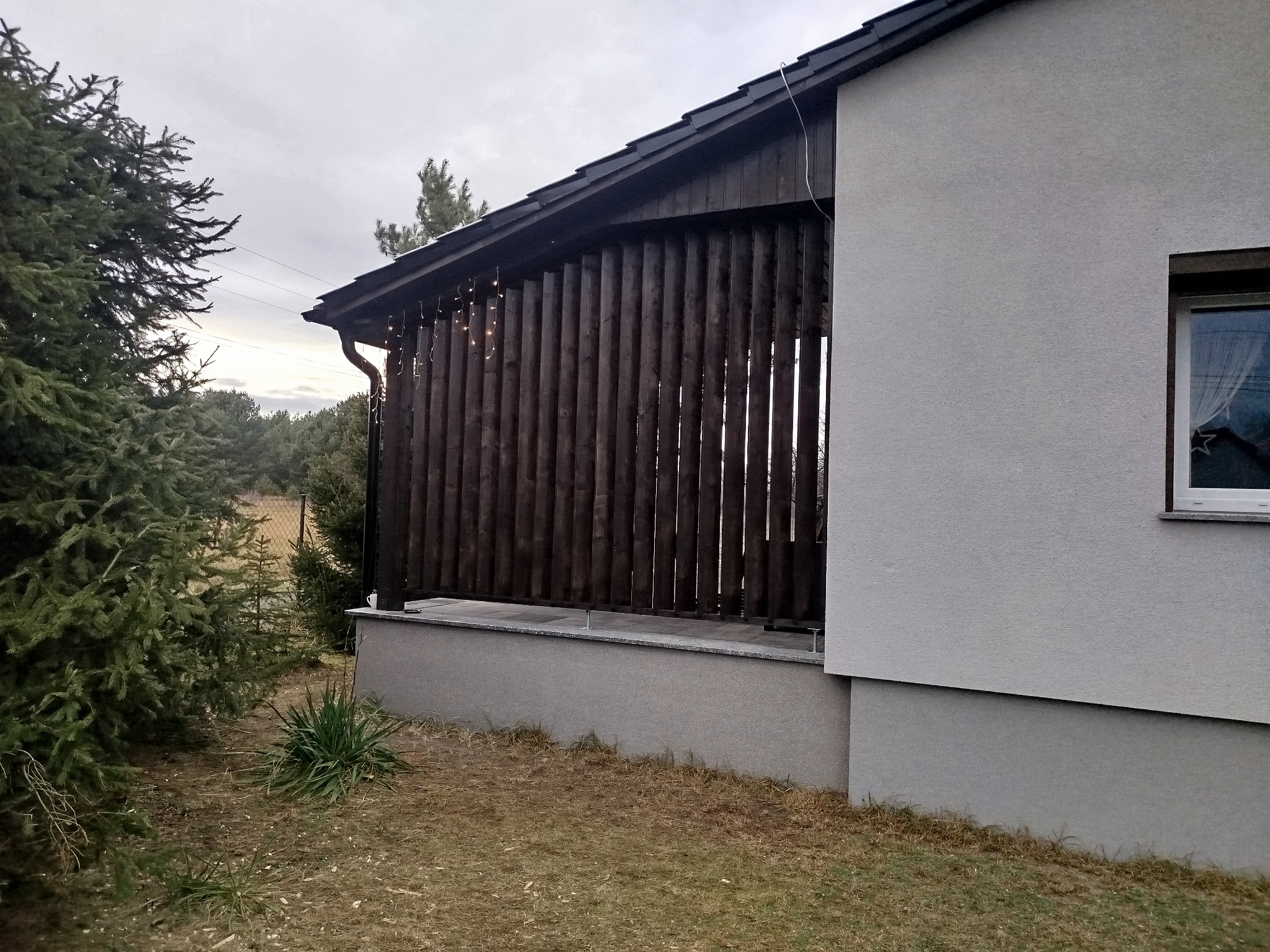 Zadaszona konstrukcja z pionowych, ciemnych desek, prawdopodobnie z oświetleniem, przylegająca do ściany domu z widocznym oknem, otoczona zielenią i drzewami iglastymi.