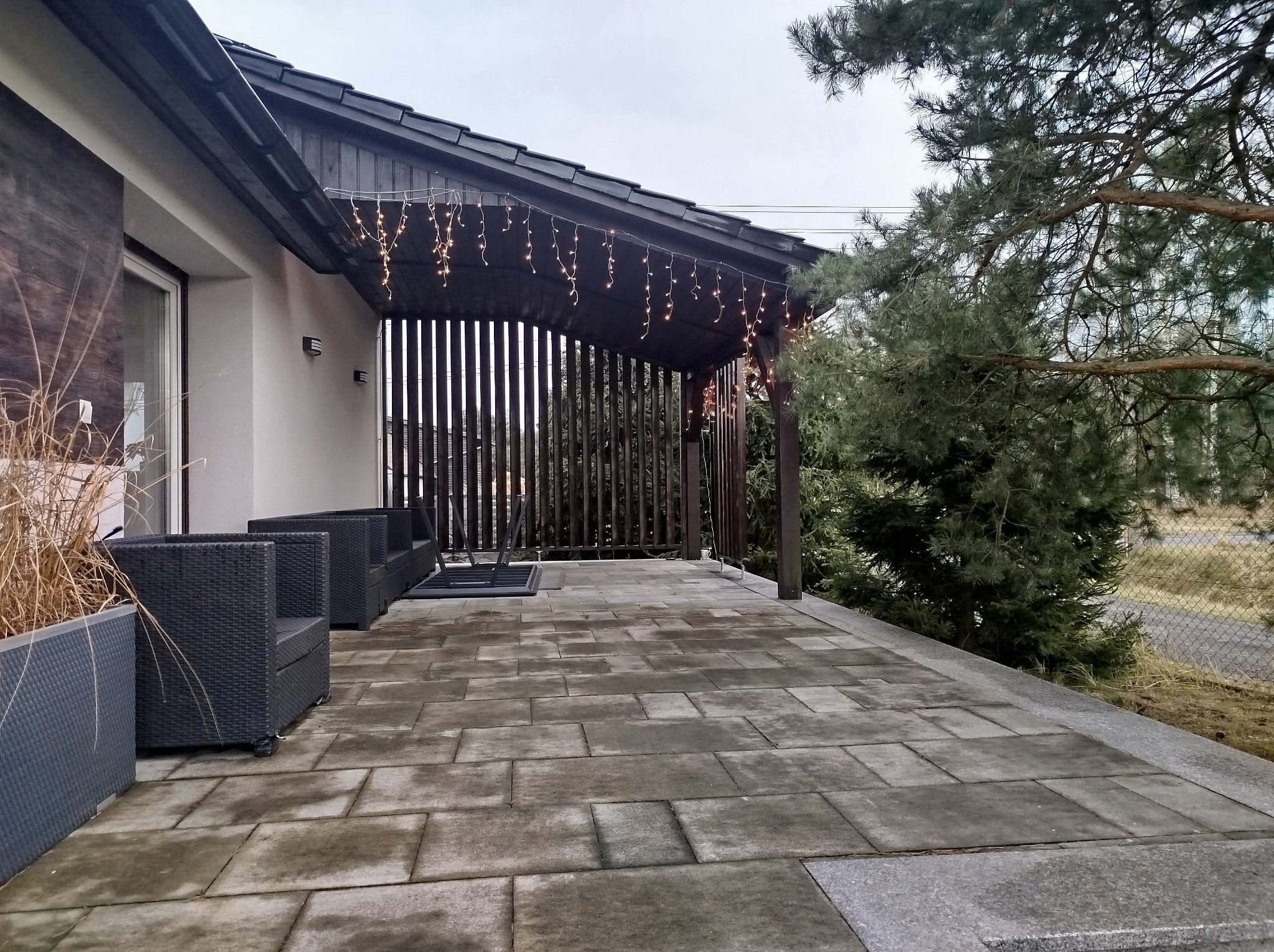 Zadaszony taras wyłożony szarą kostką brukową, z czarnymi meblami ogrodowymi i dekoracyjnym oświetleniem w postaci girlandy żarówek.