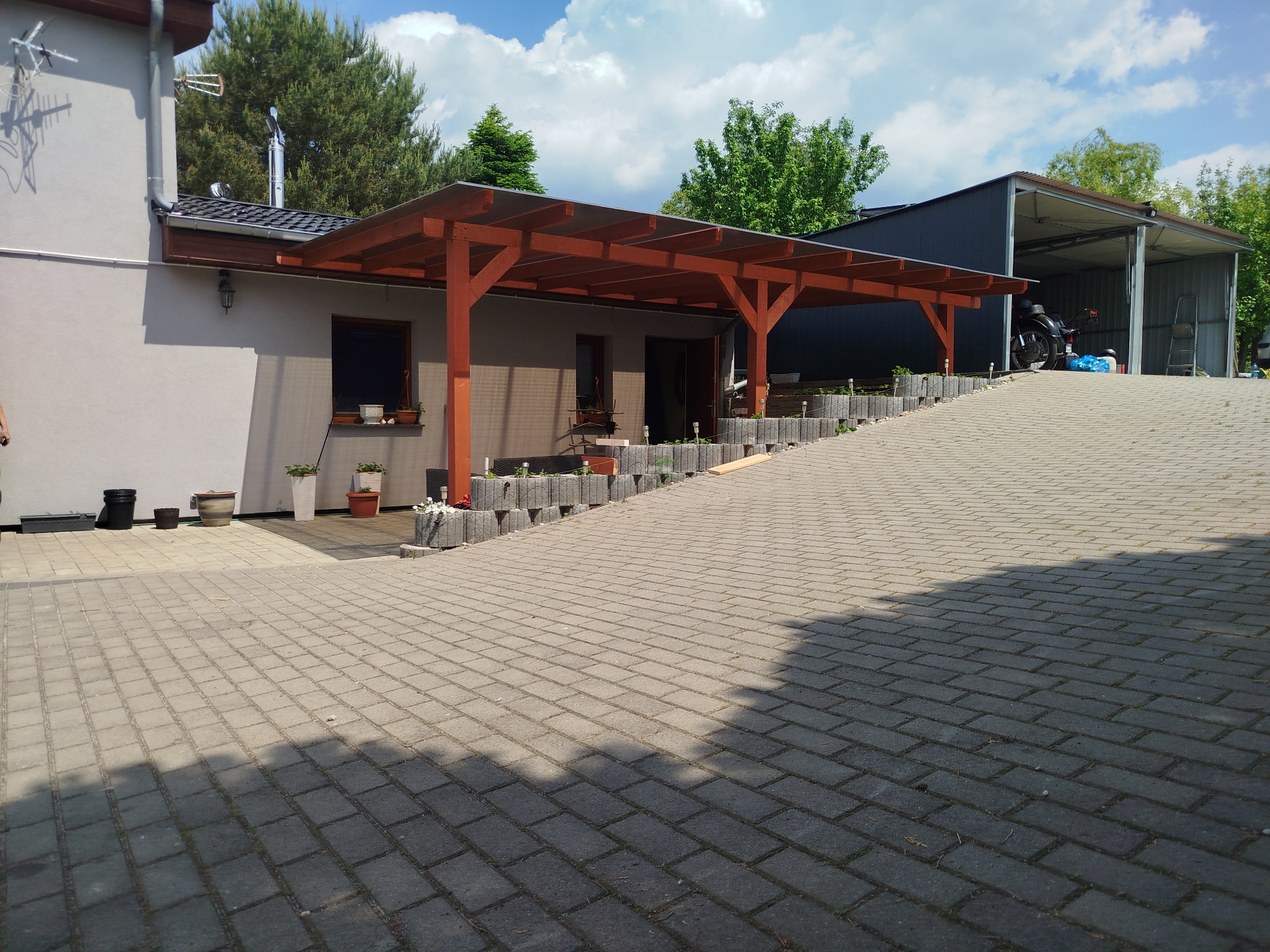 Drewniana konstrukcja z zadaszeniem przylegająca do budynku mieszkalnego, z donicami i roślinami w otoczeniu. Widok na podjazd z kostki brukowej i garaż.