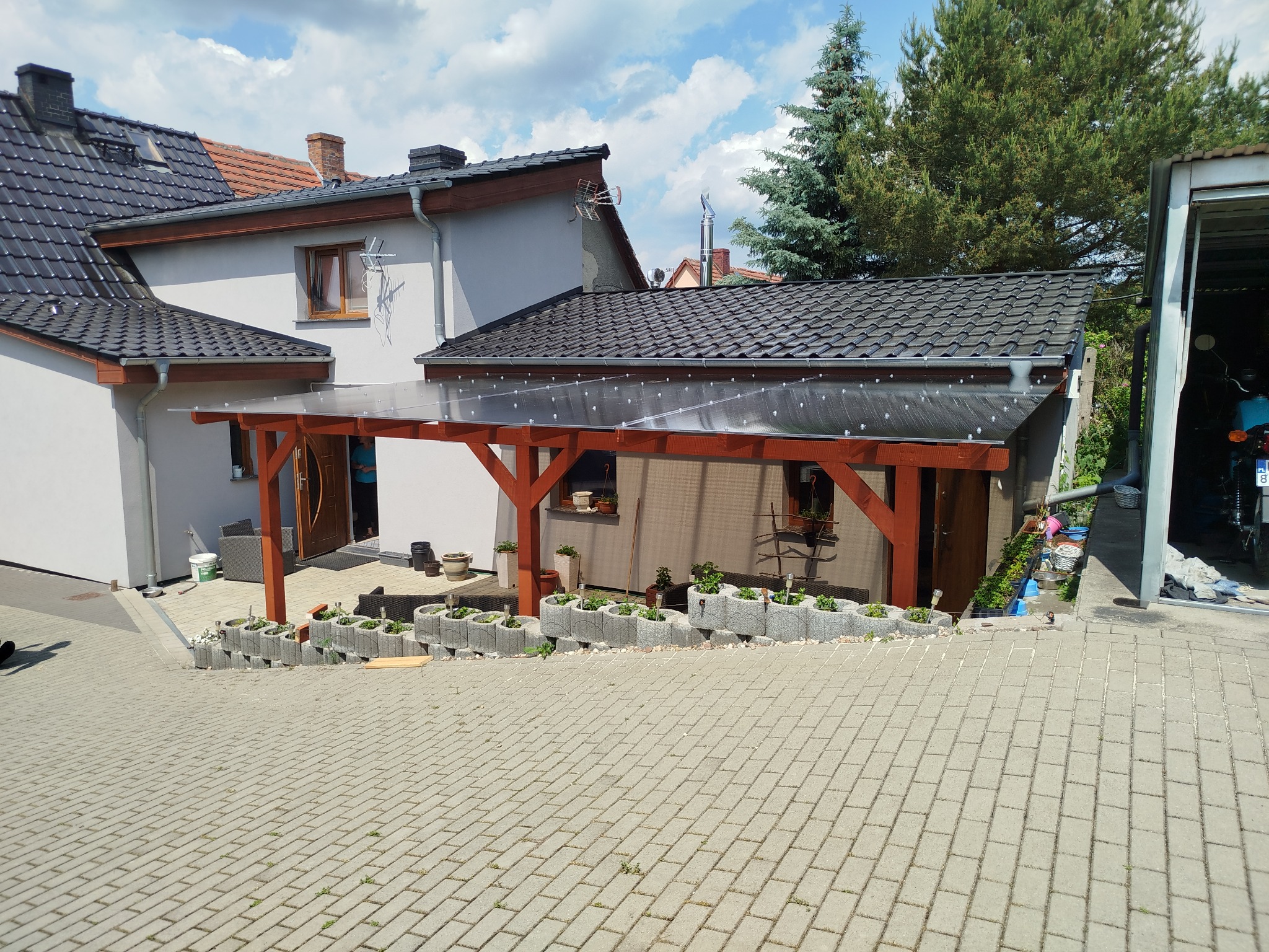 Drewniana pergola z przezroczystym zadaszeniem przy domu, z donicami na obrzeżach. Widok z brukowanej nawierzchni, w tle budynki i zieleń. Słoneczny dzień.