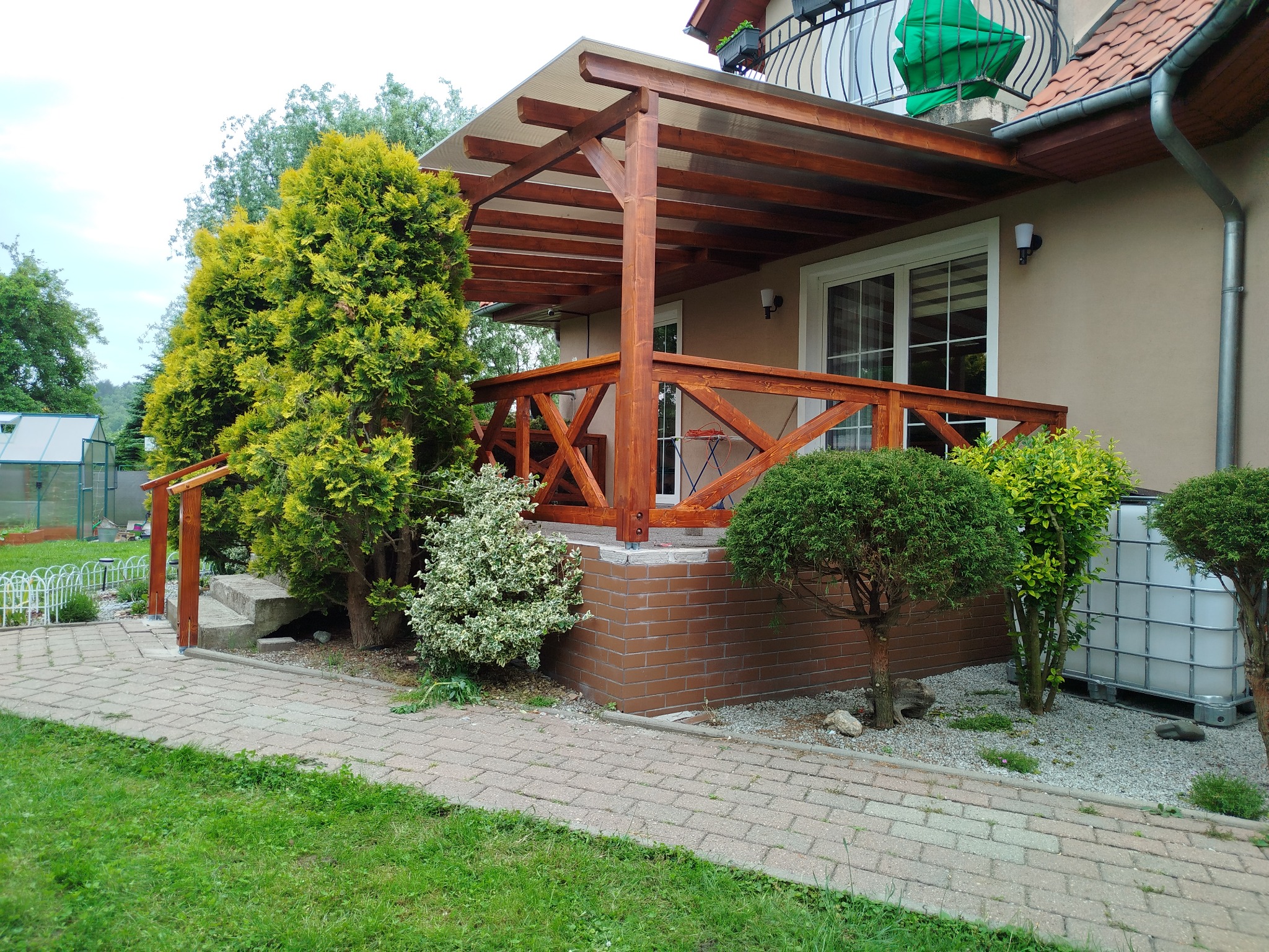 Drewniana pergola z ażurową balustradą, przylegająca do domu z balkonem. Widoczne rośliny ozdobne, ścieżka z kostki i zbiornik na deszczówkę.