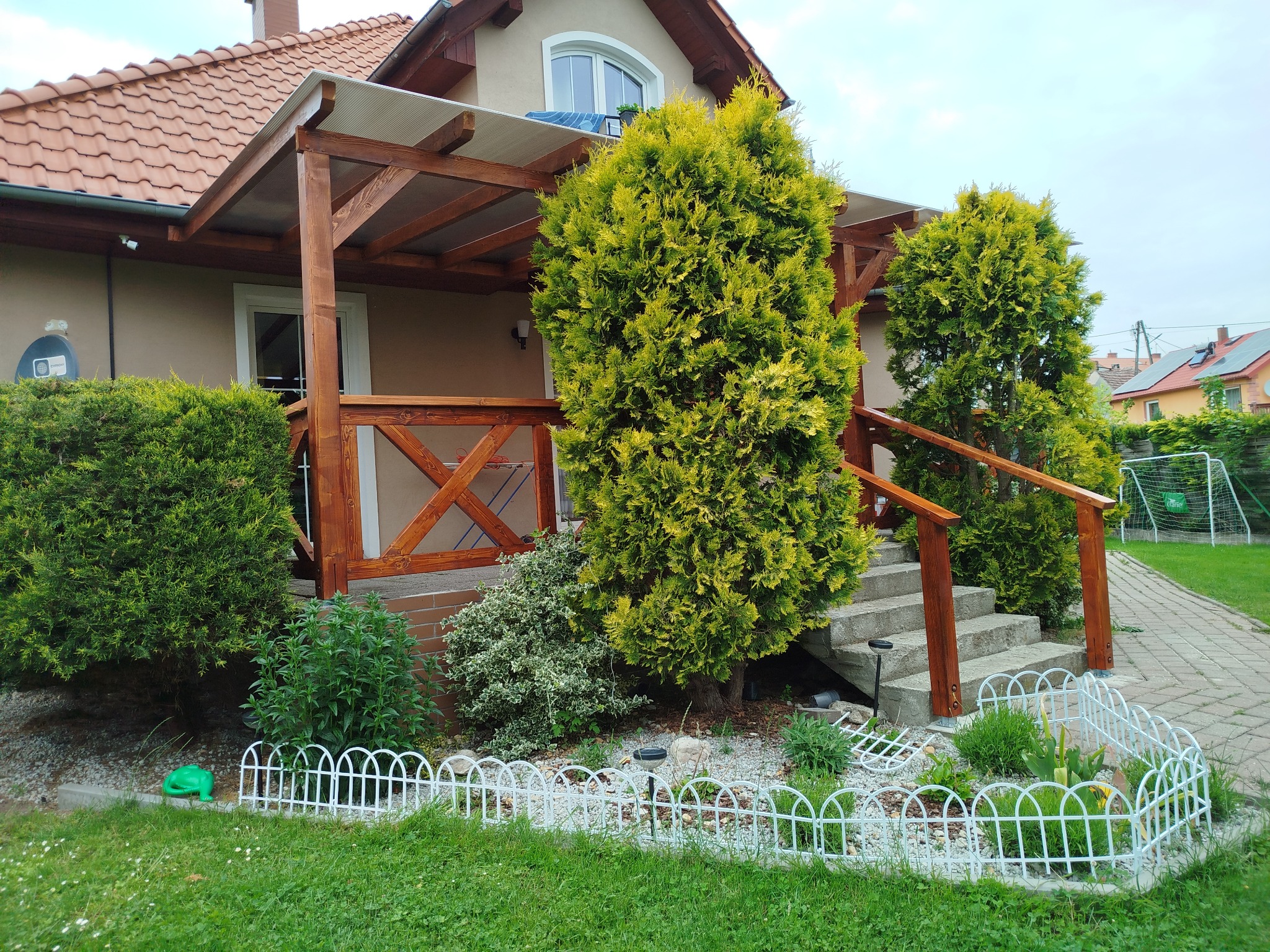 Drewniana pergola z zadaszeniem nad wejściem do domu, z ażurową balustradą i schodami. Wokół roślinność, ozdobne obrzeża rabat. Widok z ogrodu.