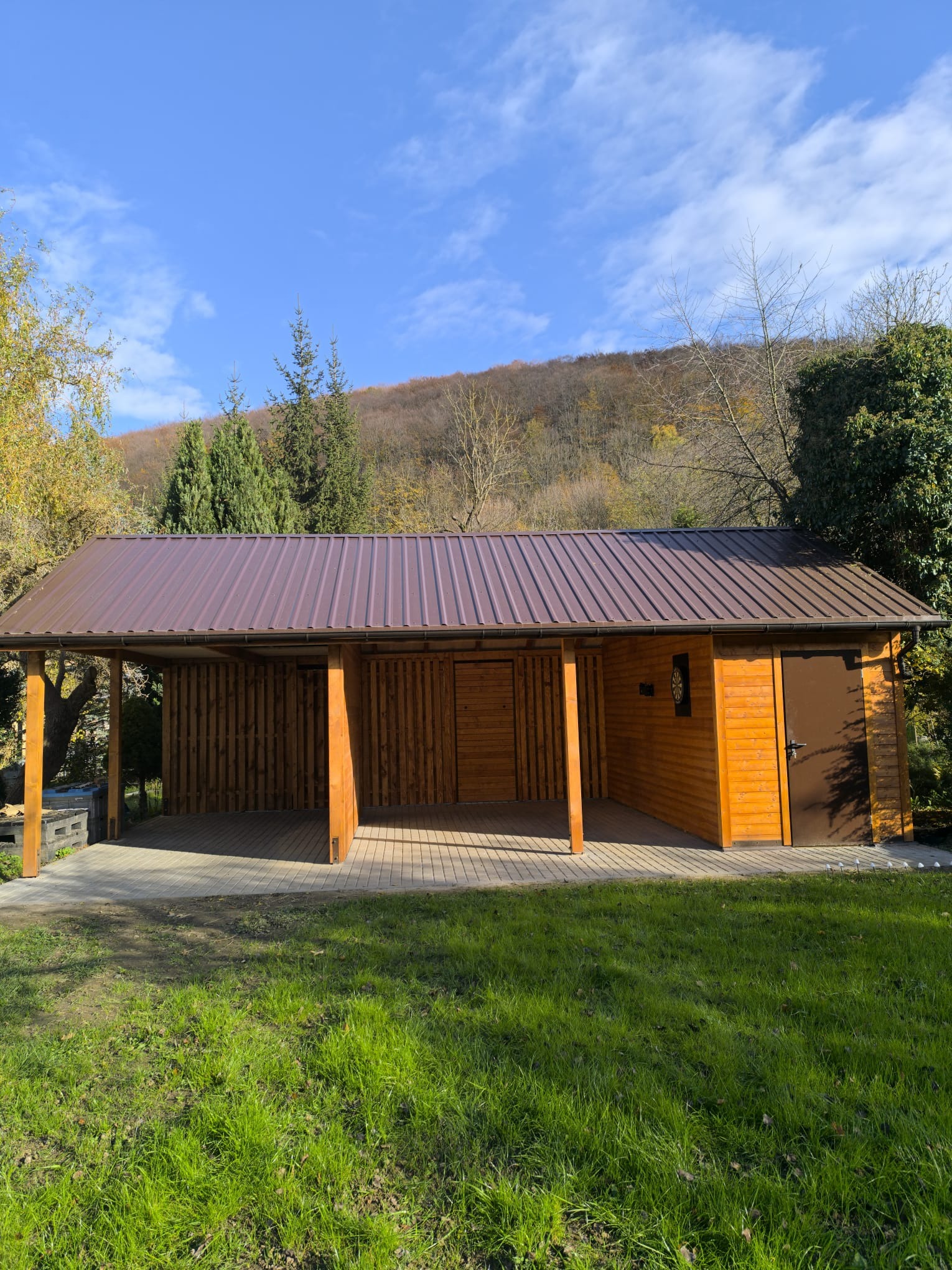 Drewniana konstrukcja z brązowym dachem na betonowej kostce, otoczona zielenią i drzewami, w tle wzgórze pod błękitnym niebem. Widoczna część zadaszona i zamknięta z drzwiami.