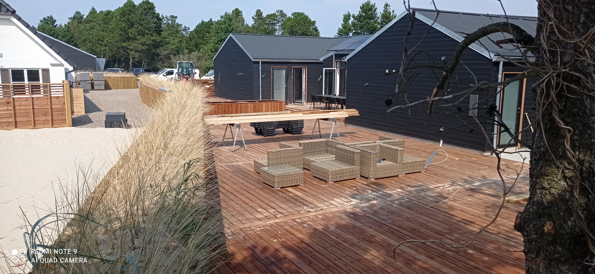 Budowa drewnianego tarasu na plaży, widoczne deski na kozłach, meble ogrodowe z rattanu i czarne domki letniskowe w tle.