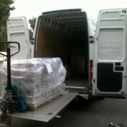 Załadunek palety owiniętej folią stretch do białego busa z otwartymi drzwiami za pomocą wózka paletowego. Widok z tyłu pojazdu, magazyn w tle.