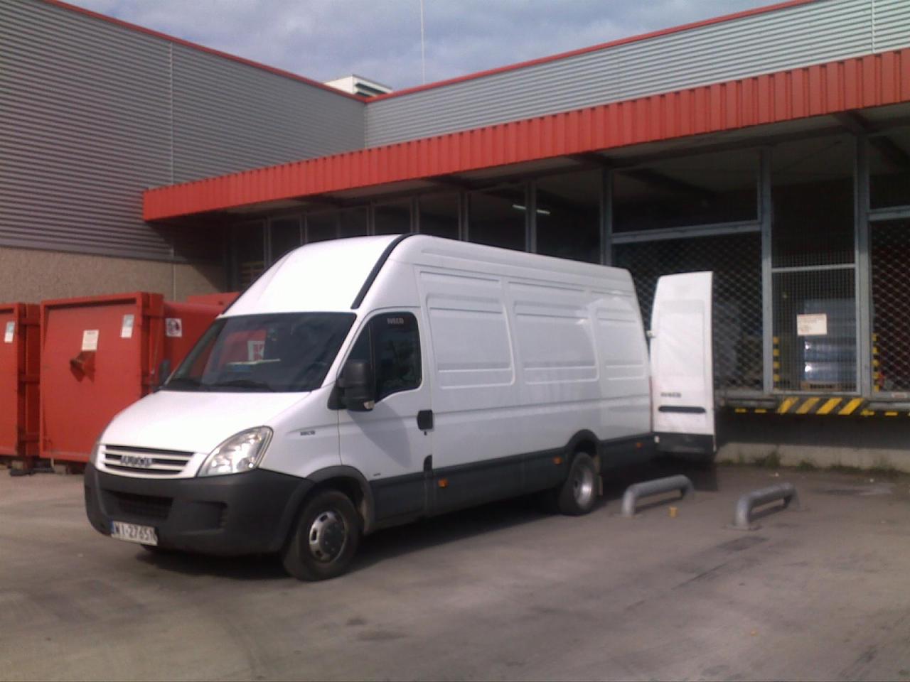 Biały bus Iveco z otwartymi tylnymi drzwiami zaparkowany na placu przed budynkiem magazynowym z czerwonymi kontenerami w tle. Widoczna tablica rejestracyjna.