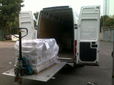 Załadunek palety owiniętej folią stretch do białego busa z otwartymi drzwiami za pomocą wózka paletowego. Widok z tyłu pojazdu, magazyn w tle.