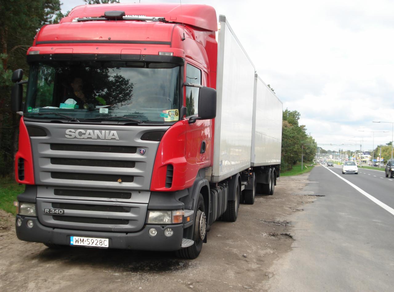 Czerwony ciągnik siodłowy Scania R340 z białą naczepą, zaparkowany na poboczu drogi. Widoczne drzewa i inne pojazdy w tle. Tablica rejestracyjna WM 5928C.