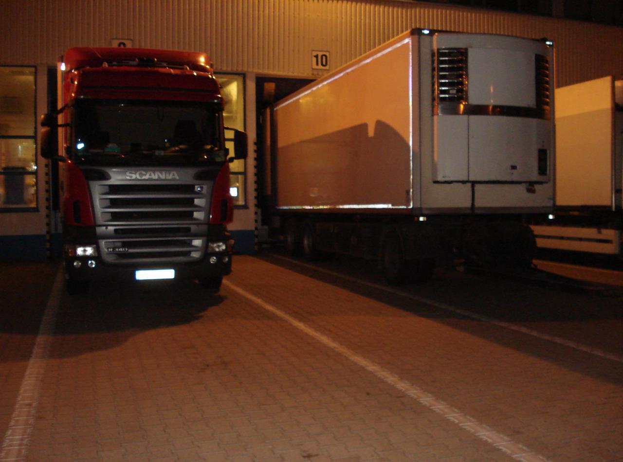 Czerwona ciężarówka Scania R 440 i biała naczepa chłodnicza zaparkowane przed magazynem z numerem 10. Oświetlenie nocne.
