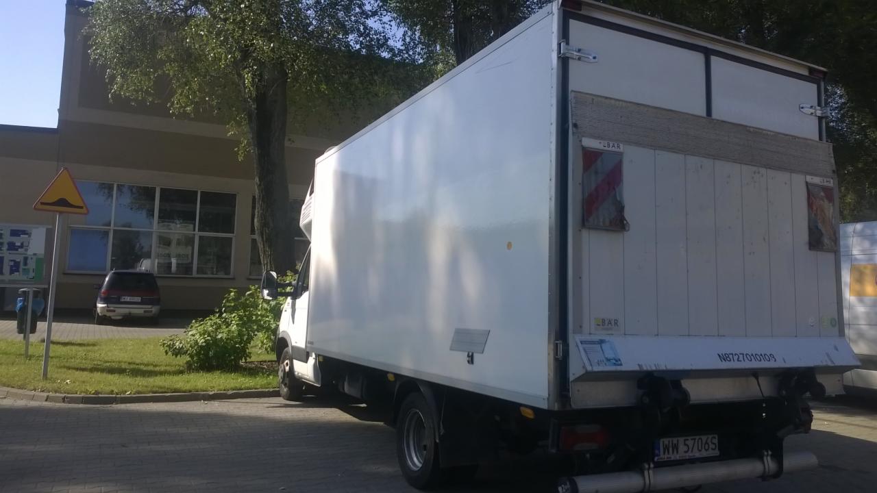 Biały samochód dostawczy z widocznymi oznaczeniami ostrzegawczymi na tylnych drzwiach, zaparkowany na zewnątrz budynku w słoneczny dzień.