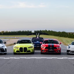 Moto Events Paweł Gmyrek - Pięć sportowych samochodów różnych marek (Toyota, BMW, Tesla, Ford Mustang, Porsche) ustawionych na linii startu toru wyścigowego, z flagami reklamowymi w tle.