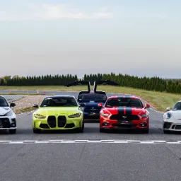 Pięć sportowych samochodów różnych marek (Toyota, BMW, Tesla, Ford Mustang, Porsche) ustawionych na linii startu toru wyścigowego, z flagami reklamowymi w tle.