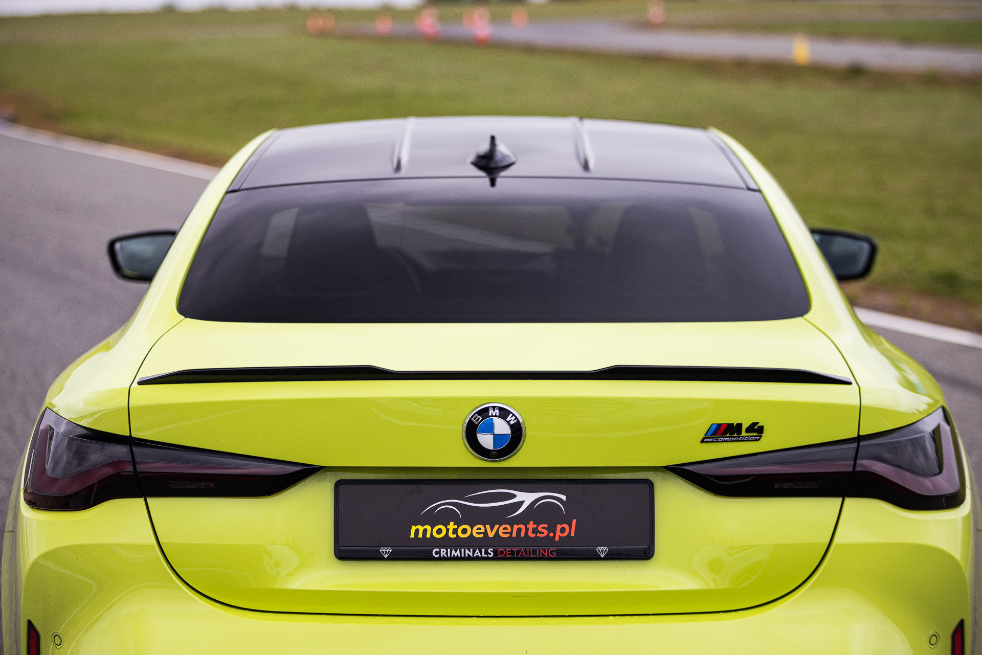 Tył cytrynowego BMW M4 Competition z czarnym dachem i logiem motoevents.pl na tablicy rejestracyjnej, stojącego na torze wyścigowym.