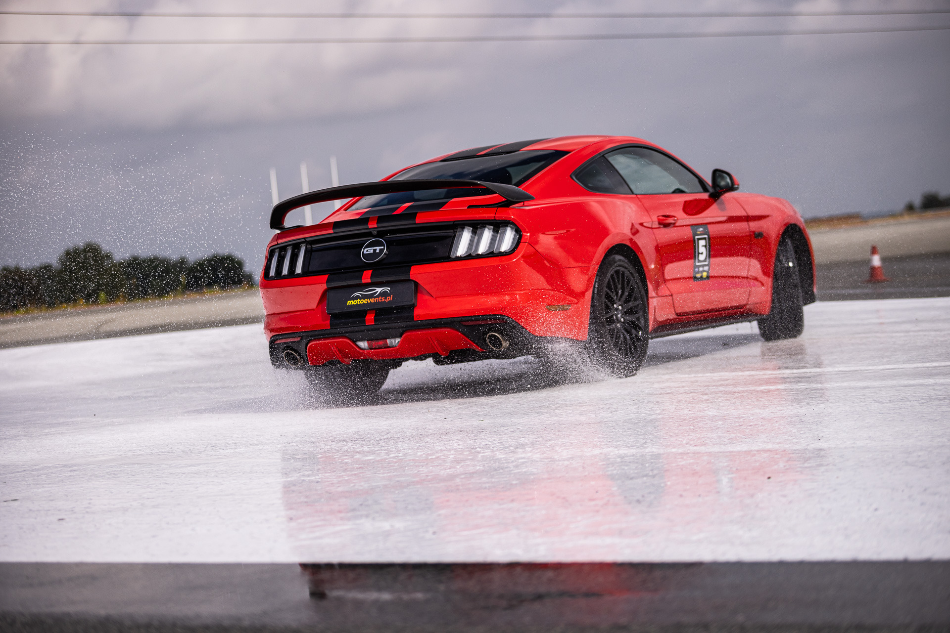 Czerwony sportowy samochód marki Ford Mustang z czarnymi pasami i numerem 5 na drzwiach, w poślizgu na mokrym torze, widok z tyłu, z logo motoevents.pl.