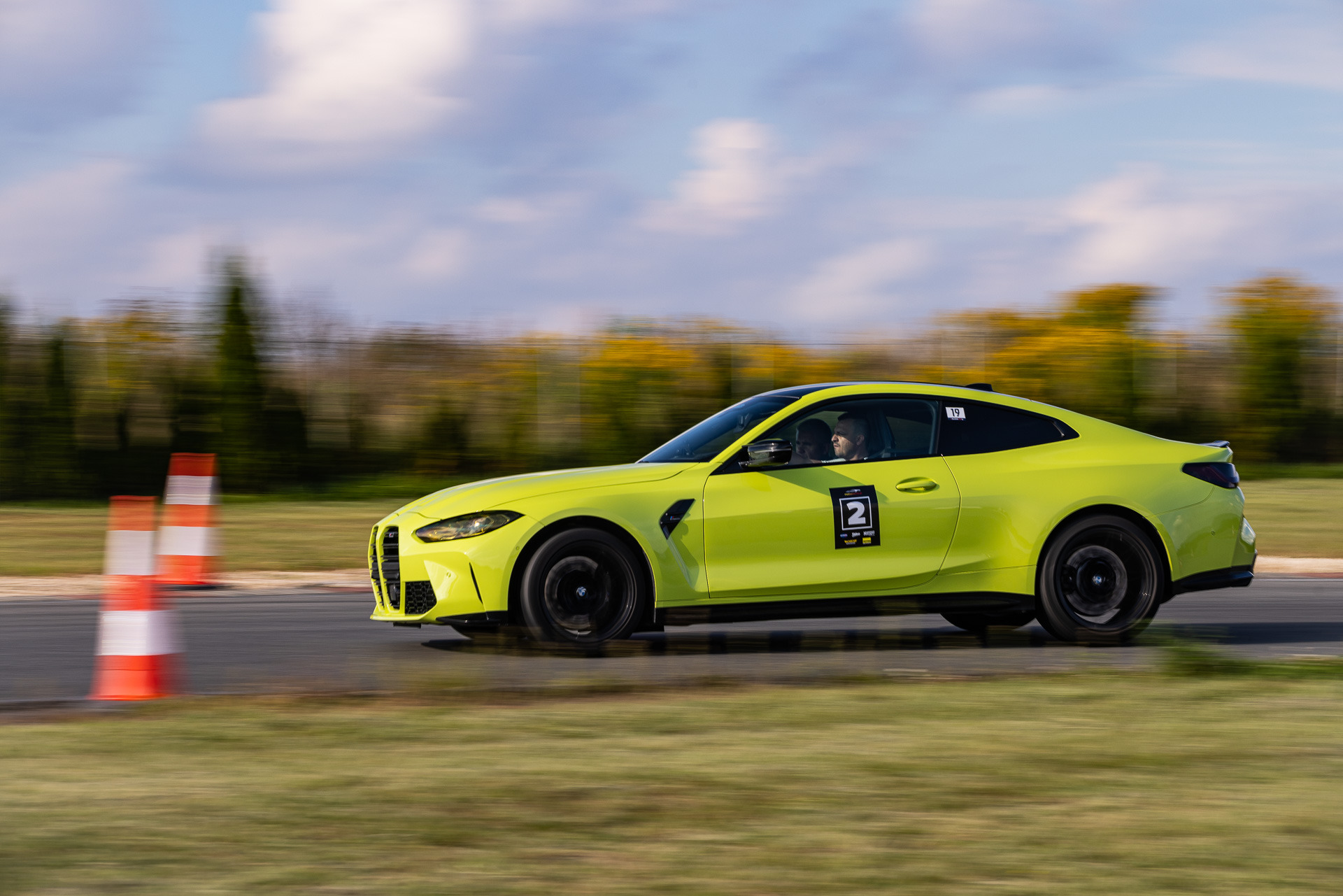 Żółty sportowy samochód BMW M4 z numerem startowym 2 na torze wyścigowym, widoczne pachołki i kierowca w kasku.