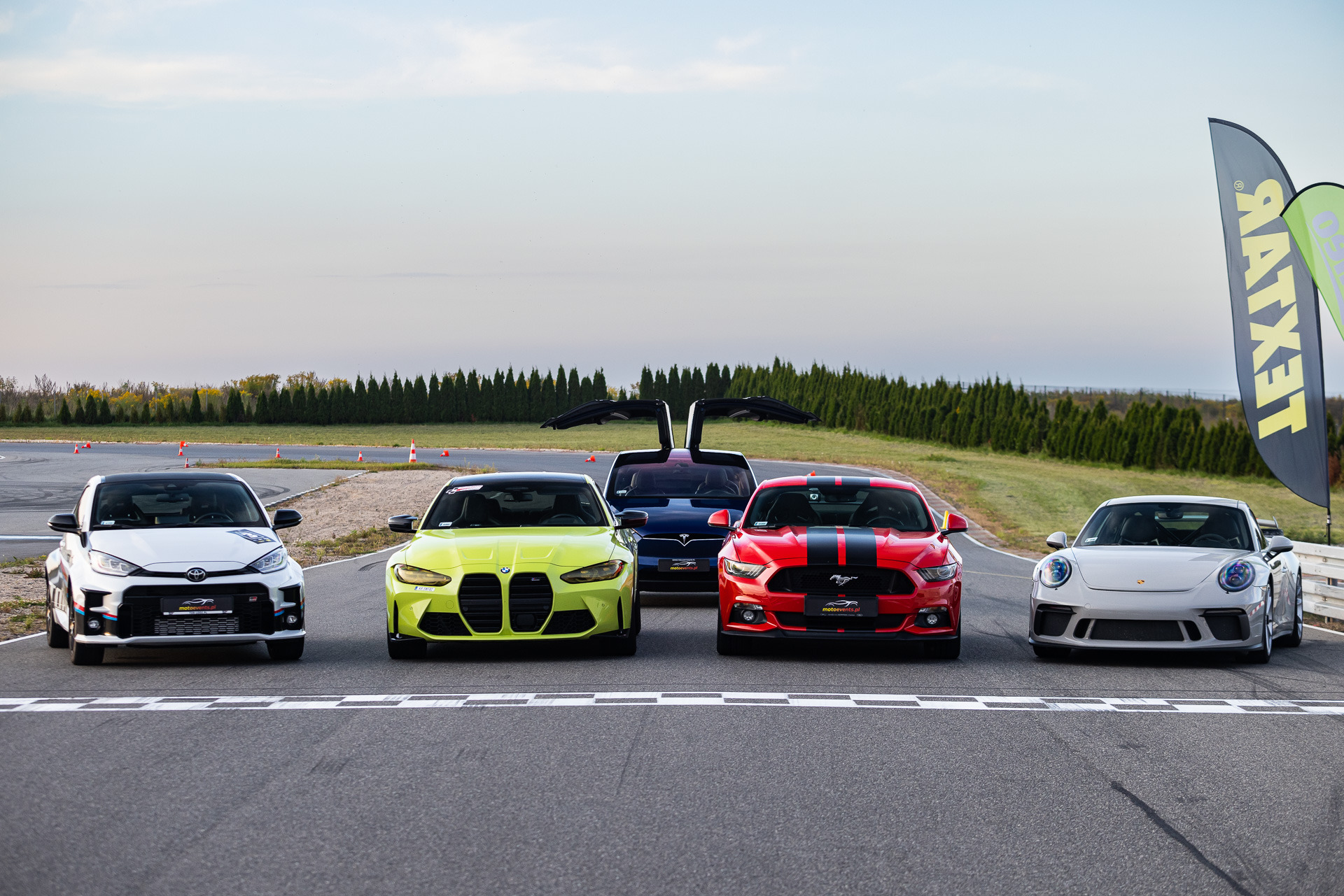 Pięć sportowych samochodów różnych marek (Toyota, BMW, Tesla, Ford Mustang, Porsche) ustawionych na linii startu toru wyścigowego, z flagami reklamowymi w tle.