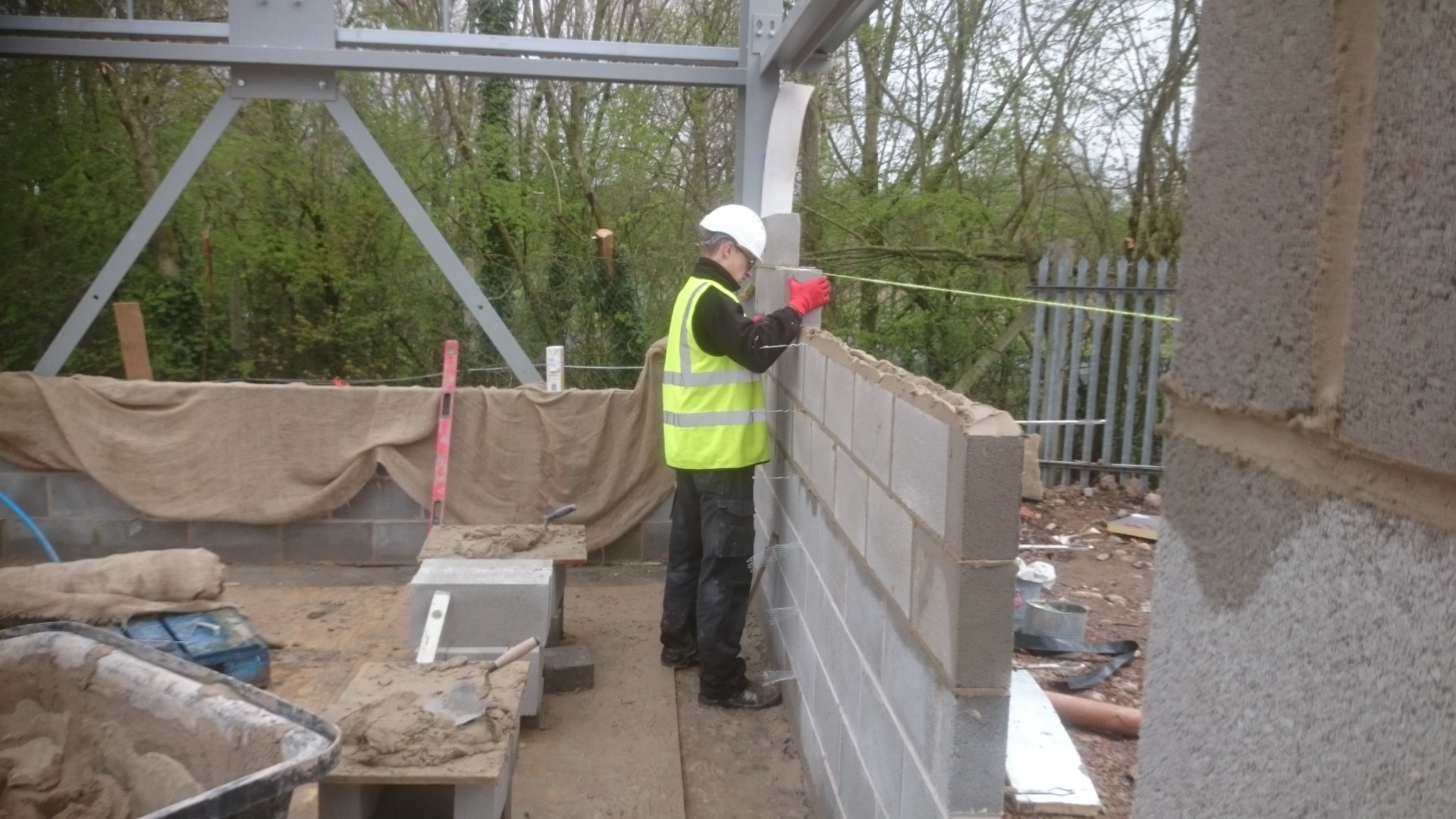 Murarz w kasku i kamizelce odblaskowej układa bloczki betonowe na powstającej ścianie budynku, widoczne stalowe elementy konstrukcyjne, zaprawa murarska i narzędzia, w tle zieleń drzew.