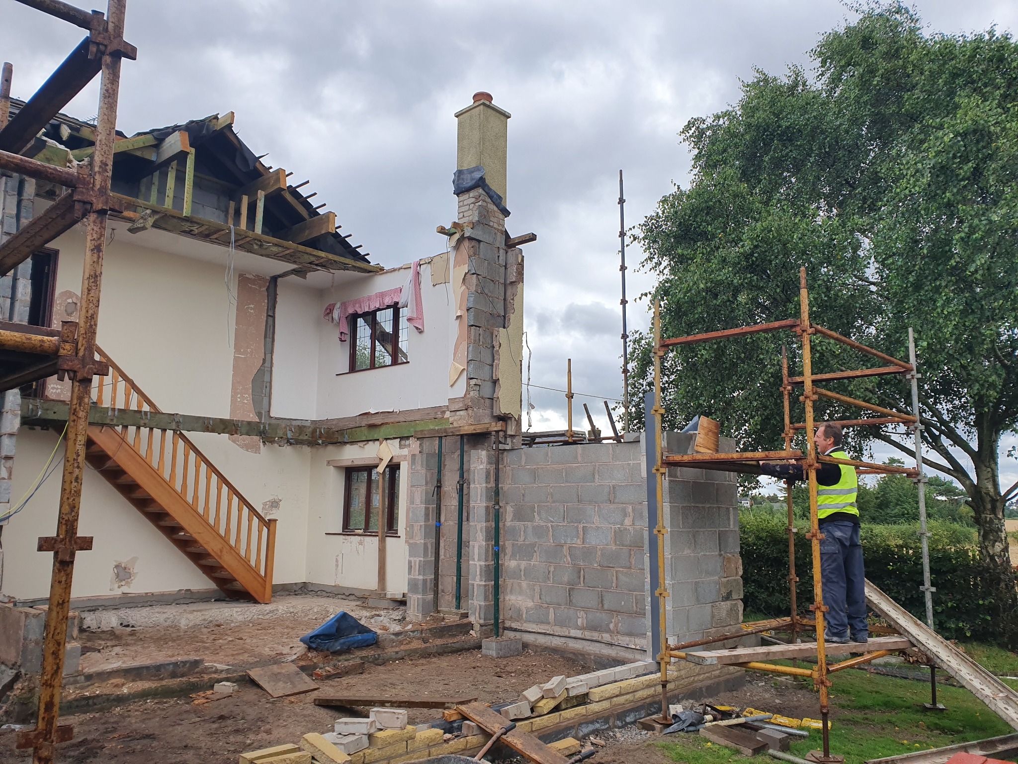 Dom w trakcie rozbudowy z widocznymi rusztowaniami, elementami konstrukcyjnymi i pracownikiem w kamizelce odblaskowej na rusztowaniu, fragmenty starej elewacji i nowy mur z bloczków betonowych.