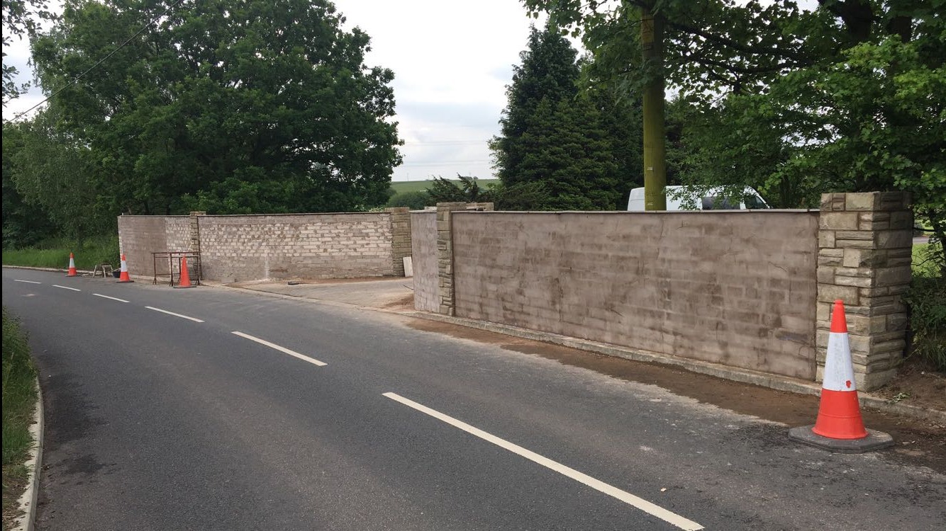 Dwa fragmenty muru oddzielone przerwą, jeden otynkowany z kamiennymi słupkami, drugi z surowej cegły, stojące przy asfalcie z pomarańczowymi pachołkami.