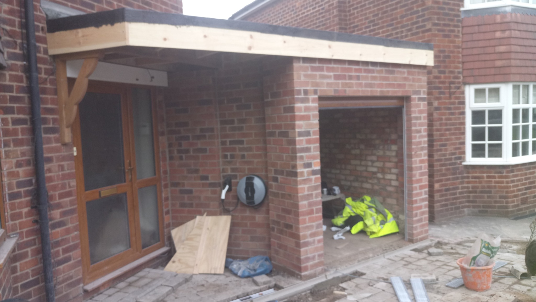 Murowana dobudówka z płaskim dachem w trakcie budowy, widoczne elementy drewniane konstrukcji dachu nad wejściem, niepodłączona ładowarka do samochodu elektrycznego na ścianie, cegła klinkierowa.