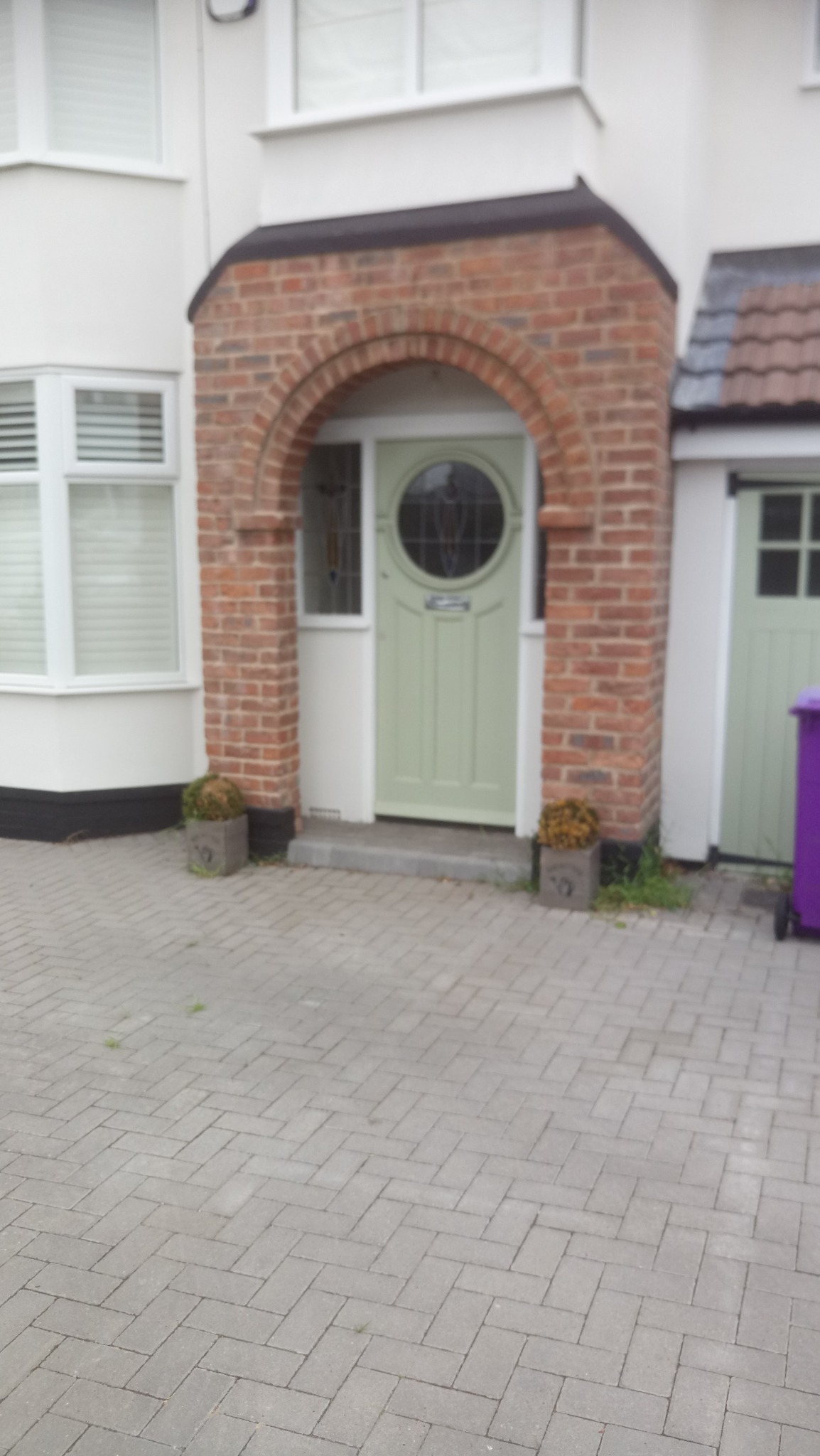 Wejście do domu z ceglanym łukiem, okrągłe okno w pastelowych zielonych drzwiach wejściowych, kostka brukowa na podjeździe.