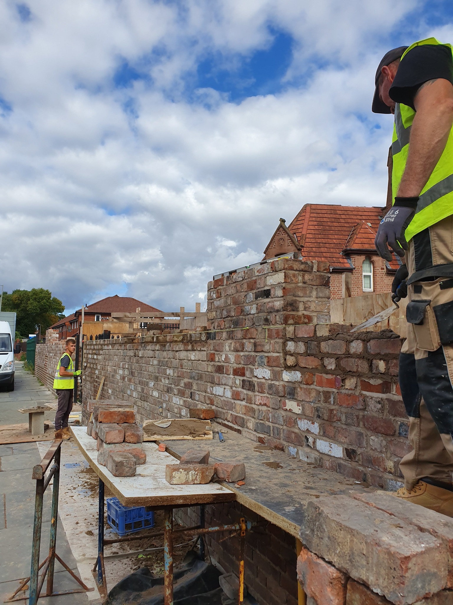 Mur z czerwonej cegły w trakcie wznoszenia, dwóch pracowników w odblaskowych kamizelkach, jeden z kielnią w ręku, drugi przy stole z cegłami, w tle zabudowa jednorodzinna i błękitne niebo.