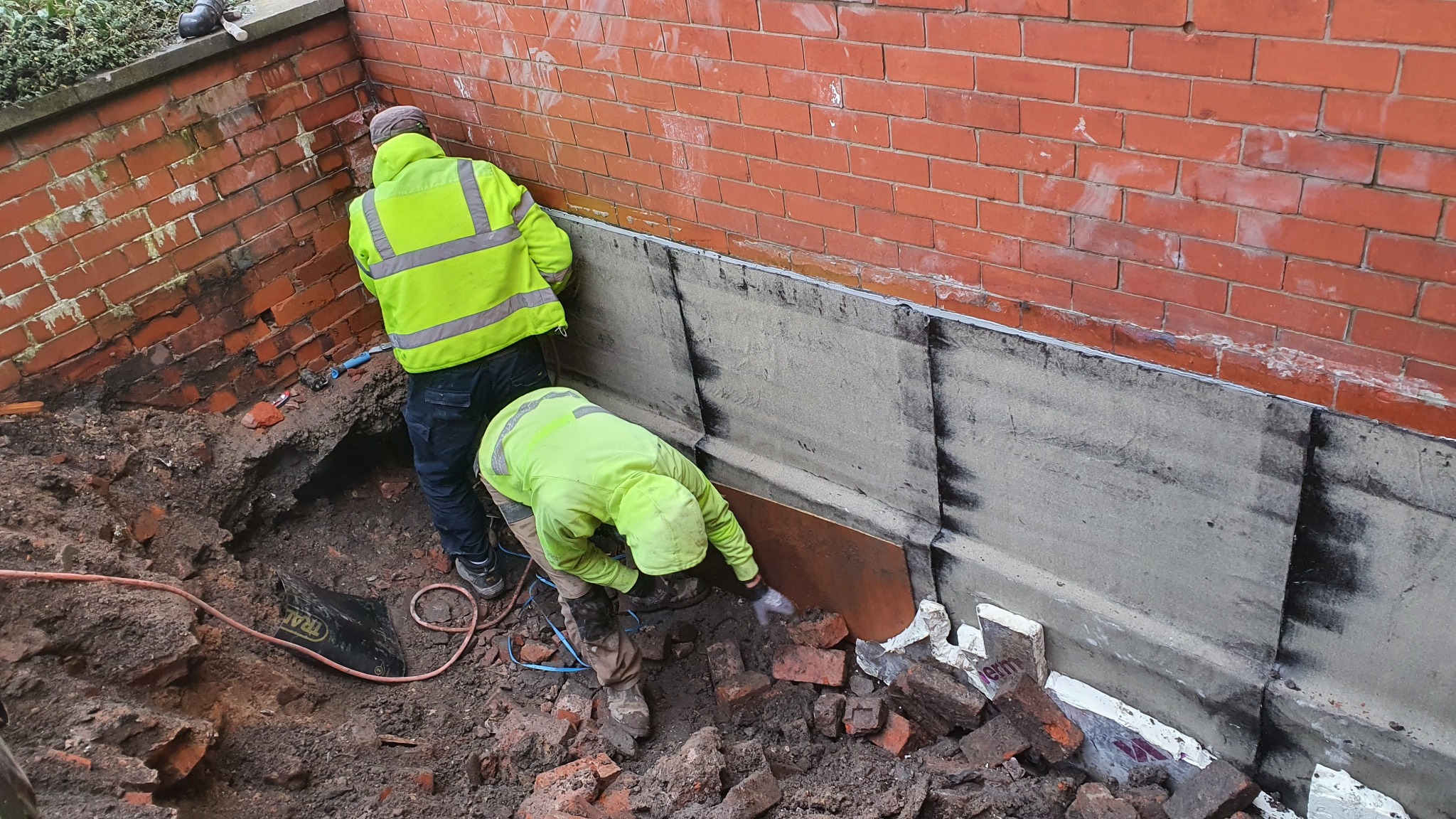 Dwóch pracowników w jaskrawych kurtkach przy ceglanym murze, jeden przykłada cegłę, drugi pracuje na górze, widoczny wykop z gruzem i narzędzia.