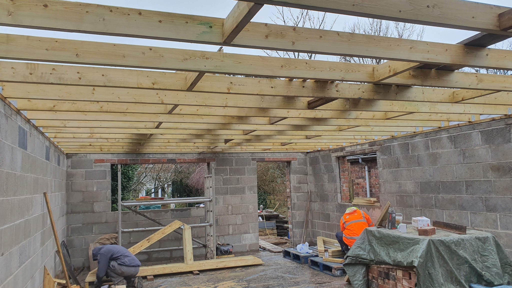 Wnętrze surowego budynku z szarych bloczków z drewnianą konstrukcją dachu w trakcie budowy, widoczni dwaj pracownicy w odzieży roboczej.