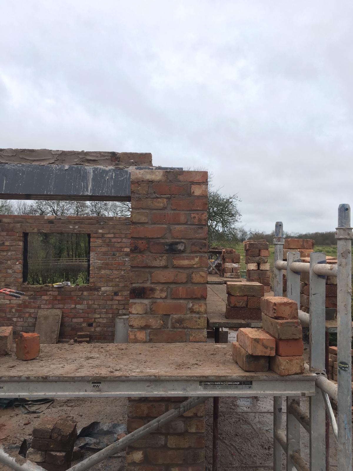 Wznoszenie muru z czerwonej cegły na budowie domu, widoczne rusztowanie i zapas cegieł, w tle zielony krajobraz pod pochmurnym niebem.