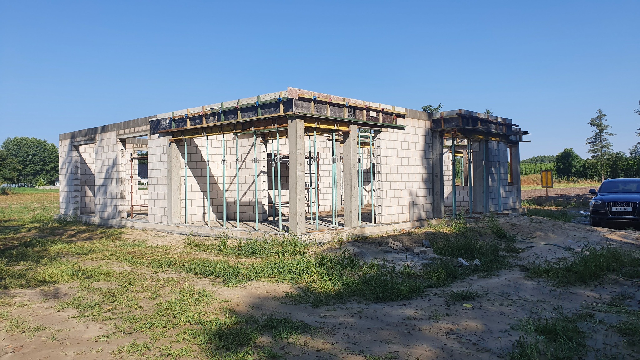 Surowy stan domu murowanego z betonu komórkowego w Liverpool, z widocznymi szalunkami i zbrojeniami, na tle błękitnego nieba i zielonej trawy. Obok zaparkowany ciemny samochód.