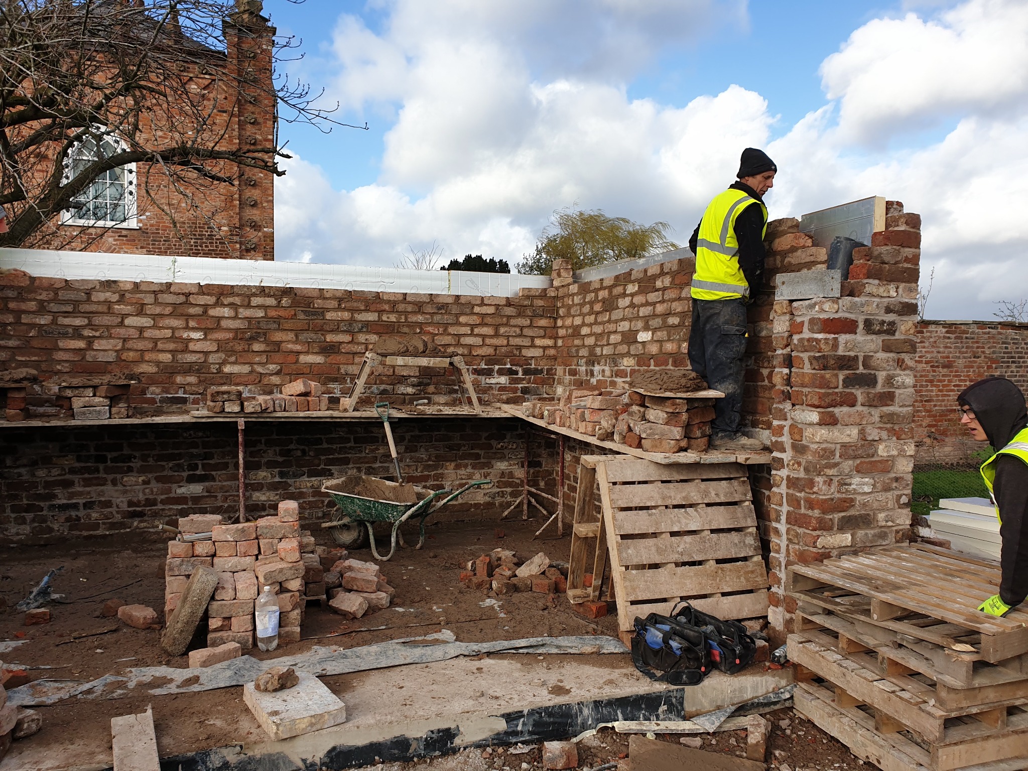 Murarz w żółtej kamizelce układa cegły na wznoszonej ścianie budynku z cegły, widoczny fragment starego budynku w tle, palety, taczka i narzędzia budowlane.