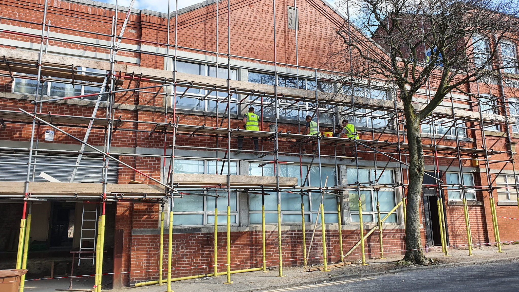 Trzech pracowników w jaskrawych kamizelkach na rusztowaniu przy murowanym budynku z licznymi oknami, widoczny fragment nagiej gałęzi drzewa po prawej stronie.