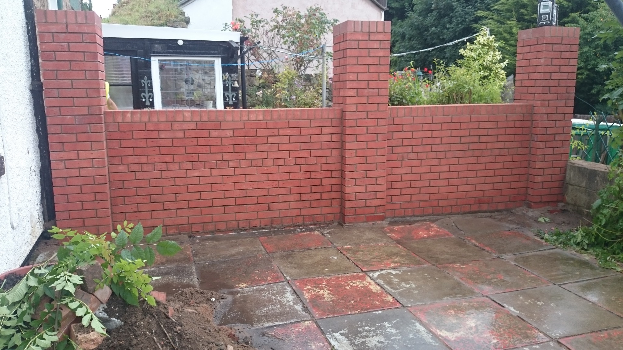Czerwony mur z cegły z filarami, oddzielający posesję, widok na fragment posesji sąsiada i element budynku