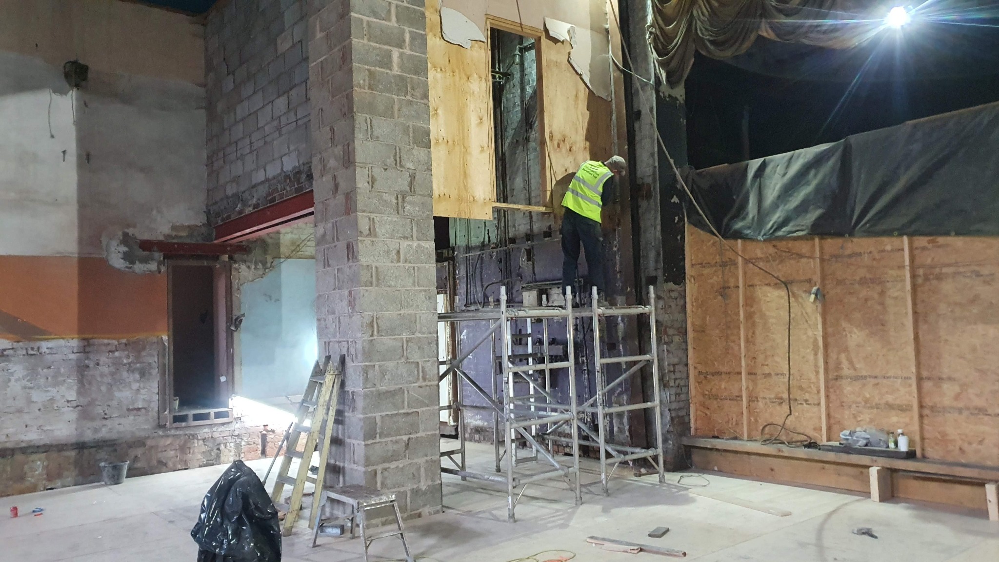 Pracownik w kamizelce odblaskowej na rusztowaniu w trakcie remontu wnętrza budynku z odsłoniętą cegłą, fragmentami płyt drewnianych i widocznymi instalacjami.