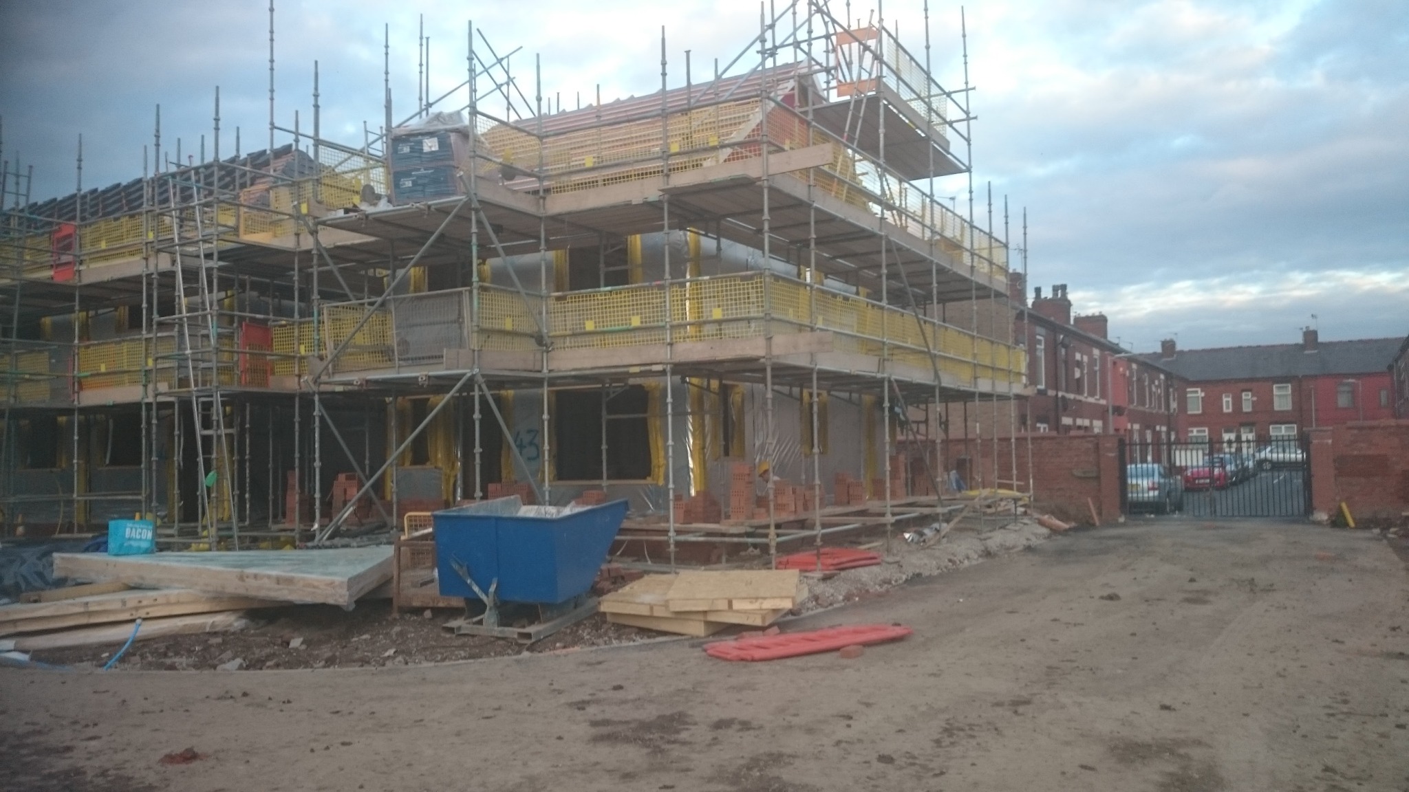 Konstrukcja domu w trakcie budowy z rusztowaniami, widoczne cegły i materiały budowlane, w tle zabudowa miejska i pochmurne niebo.