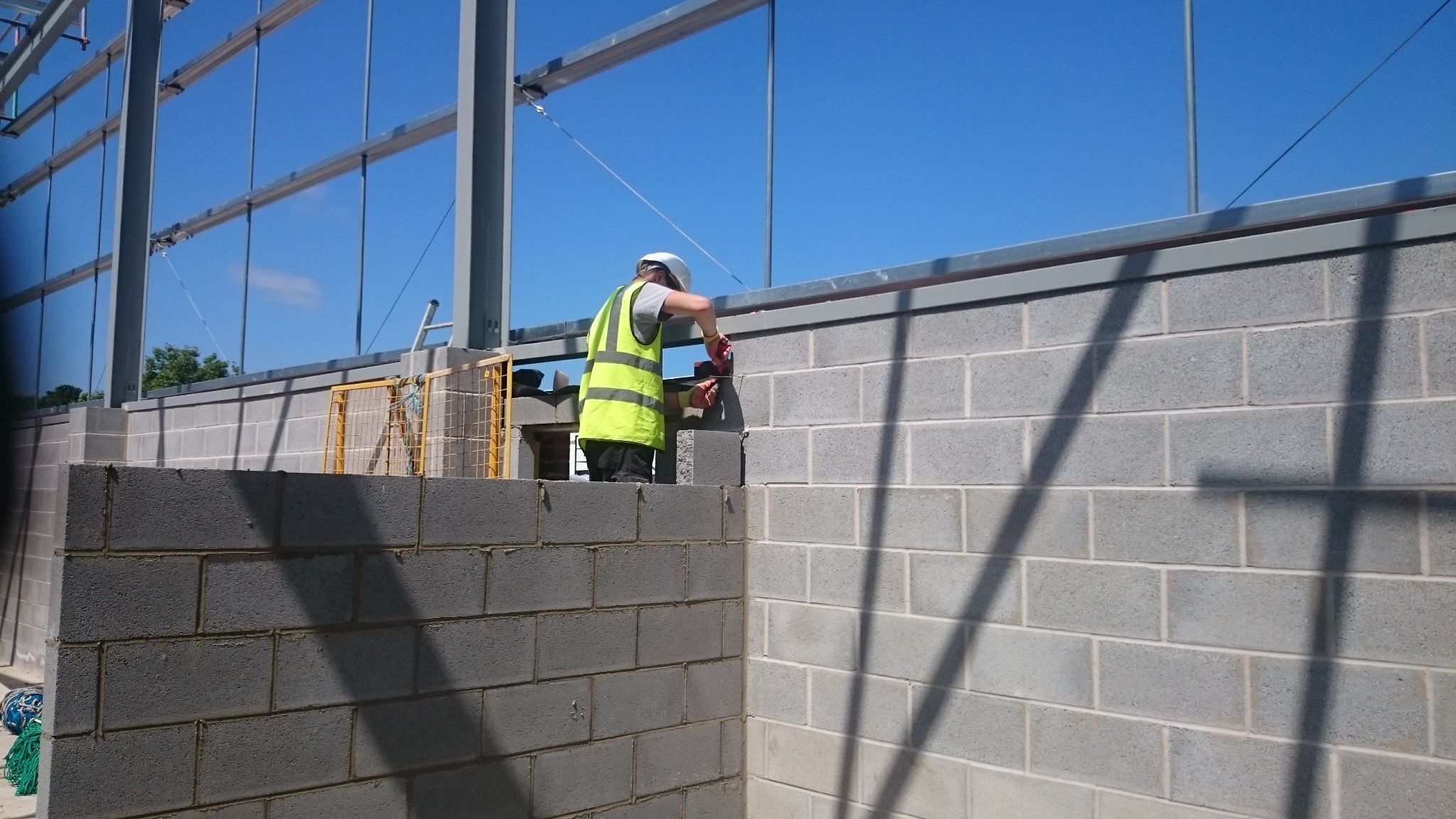 Pracownik w kasku i kamizelce odblaskowej muruje ścianę z szarych bloczków betonowych na tle konstrukcji stalowej i błękitnego nieba.