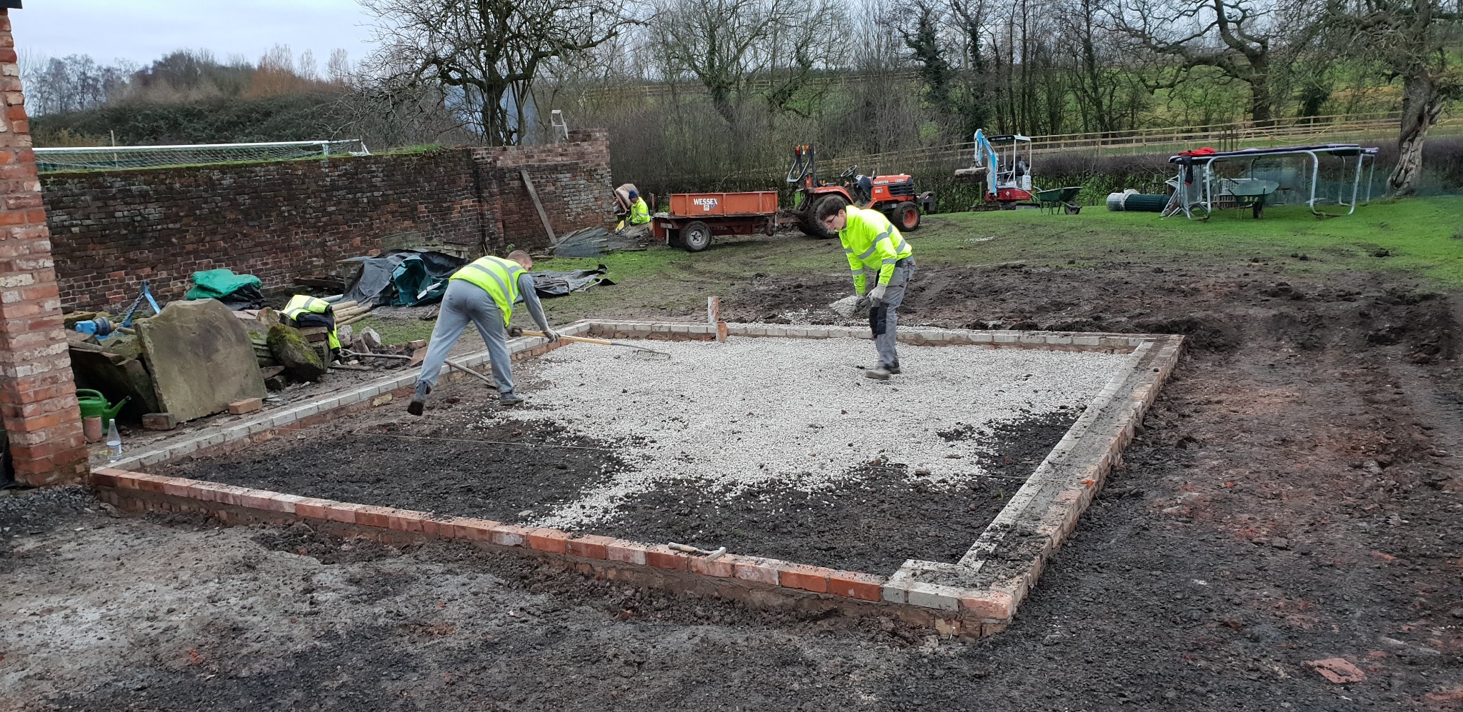 Budowa fundamentów pod dom: robotnicy w kamizelkach odblaskowych rozsypują żwir na przygotowanym terenie z ceglaną obwódką, w tle sprzęt budowlany i drzewa.