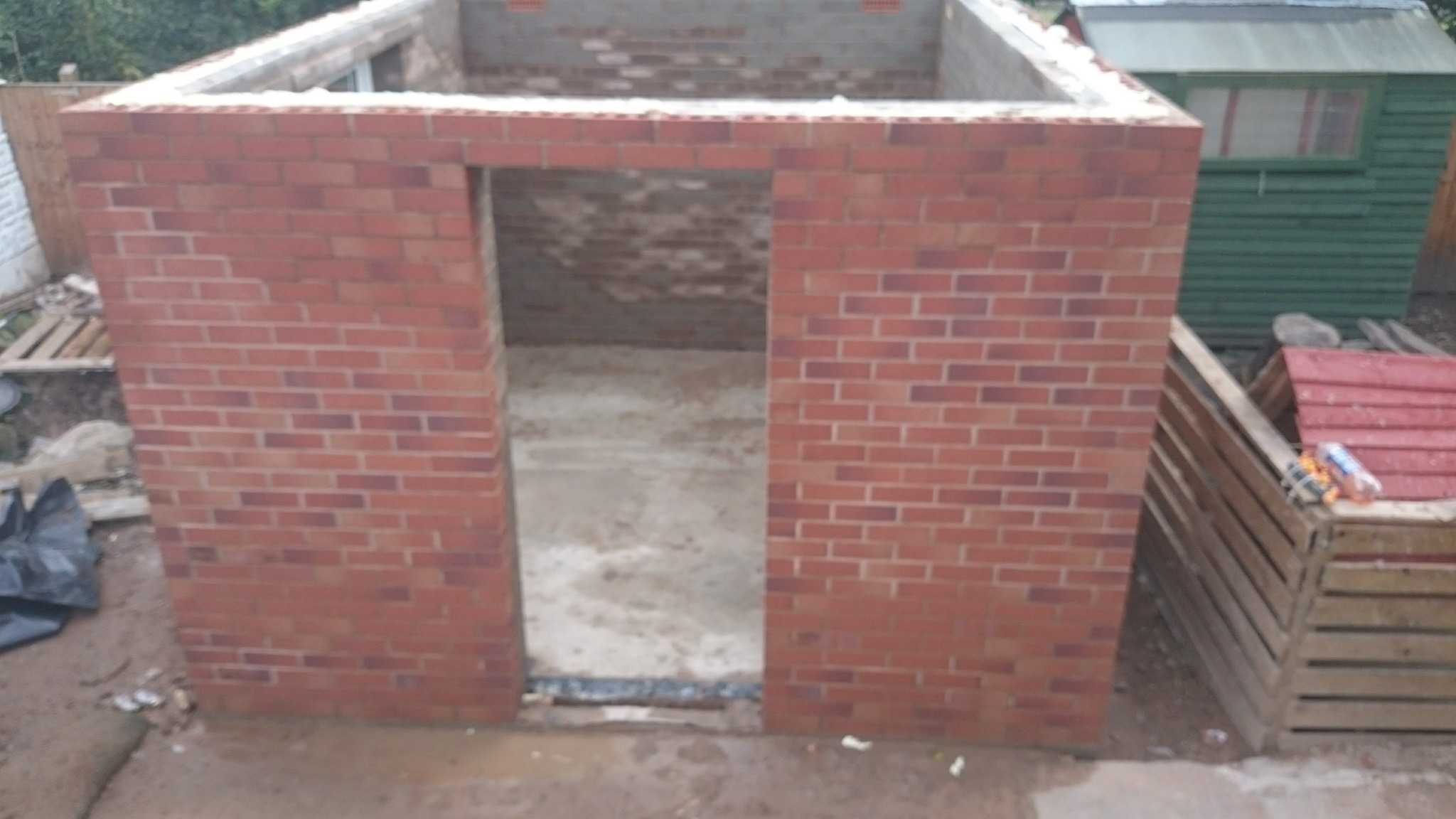 Surowa konstrukcja ścian z czerwonej cegły z otworem drzwiowym, widoczny betonowy fundament wewnątrz, w tle zielony budynek gospodarczy i palety.