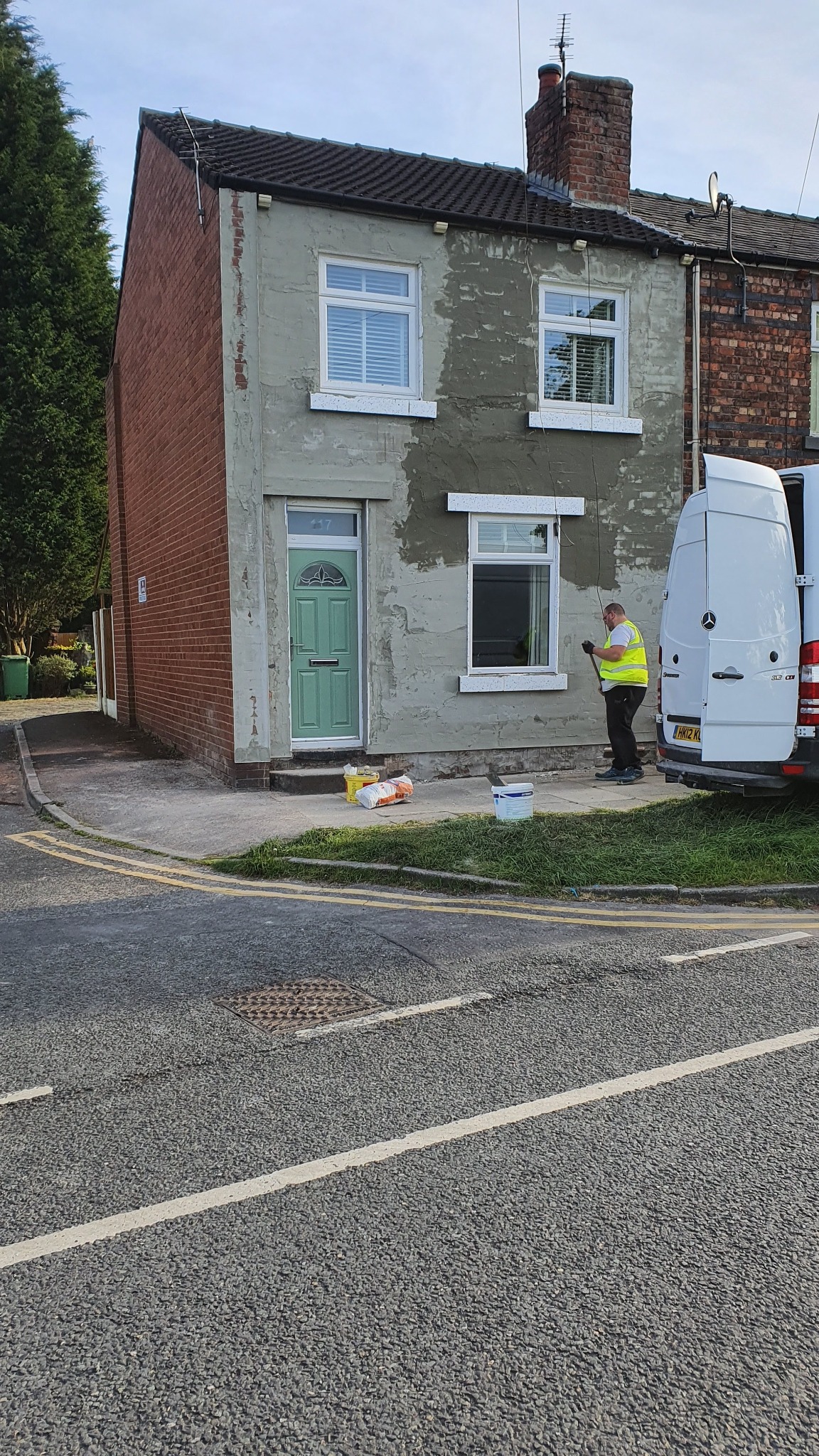 Pracownik w kamizelce odblaskowej maluje elewację domu jednorodzinnego na szaro, widoczny fragment ceglanej ściany bocznej, białe ramy okienne i zielone drzwi wejściowe, obok stoi biały samochód...