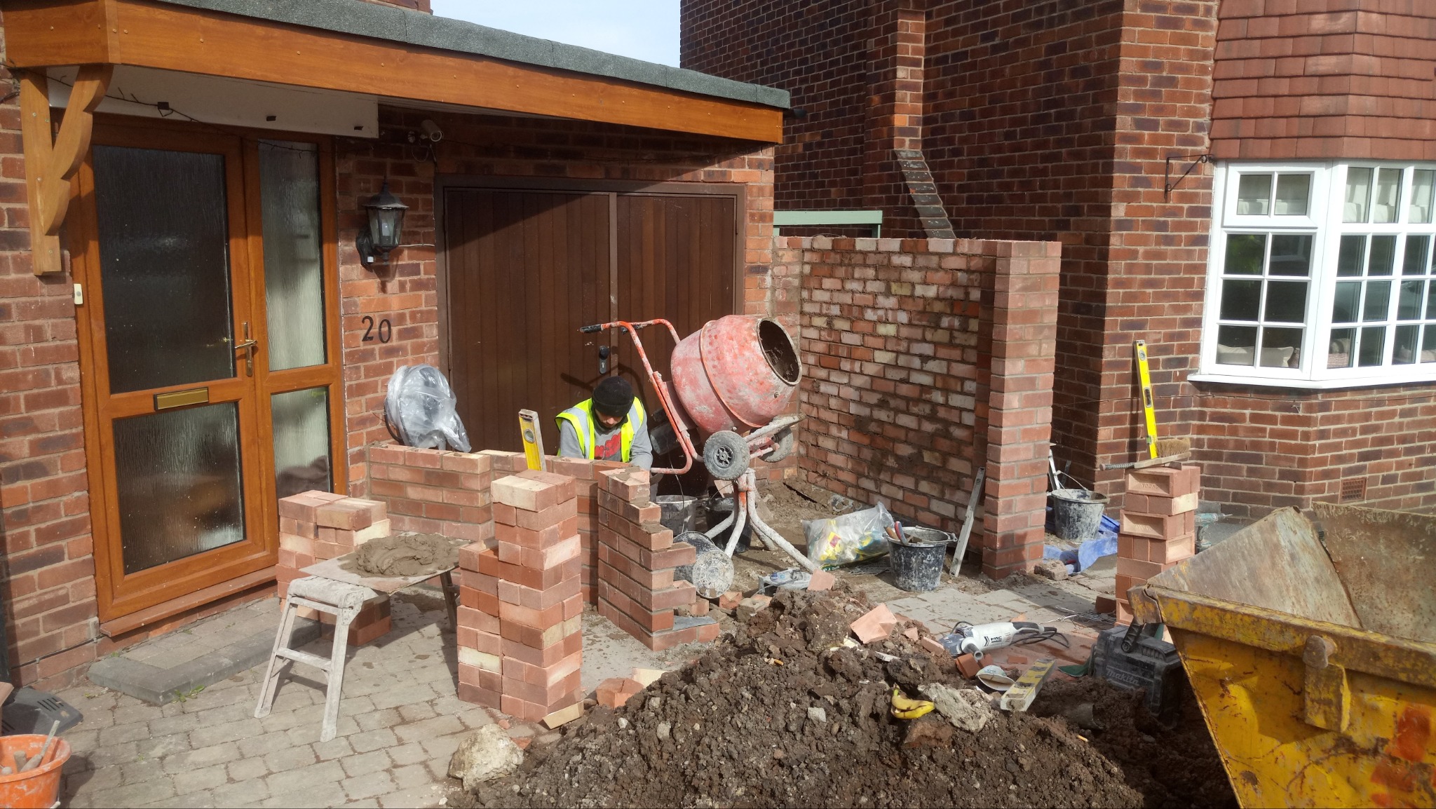 Prace murarskie przed domem z czerwonej cegły: murarz w kamizelce odblaskowej układa cegły, obok stoi brudna betoniarka, widoczny kontener na gruz i narzędzia.