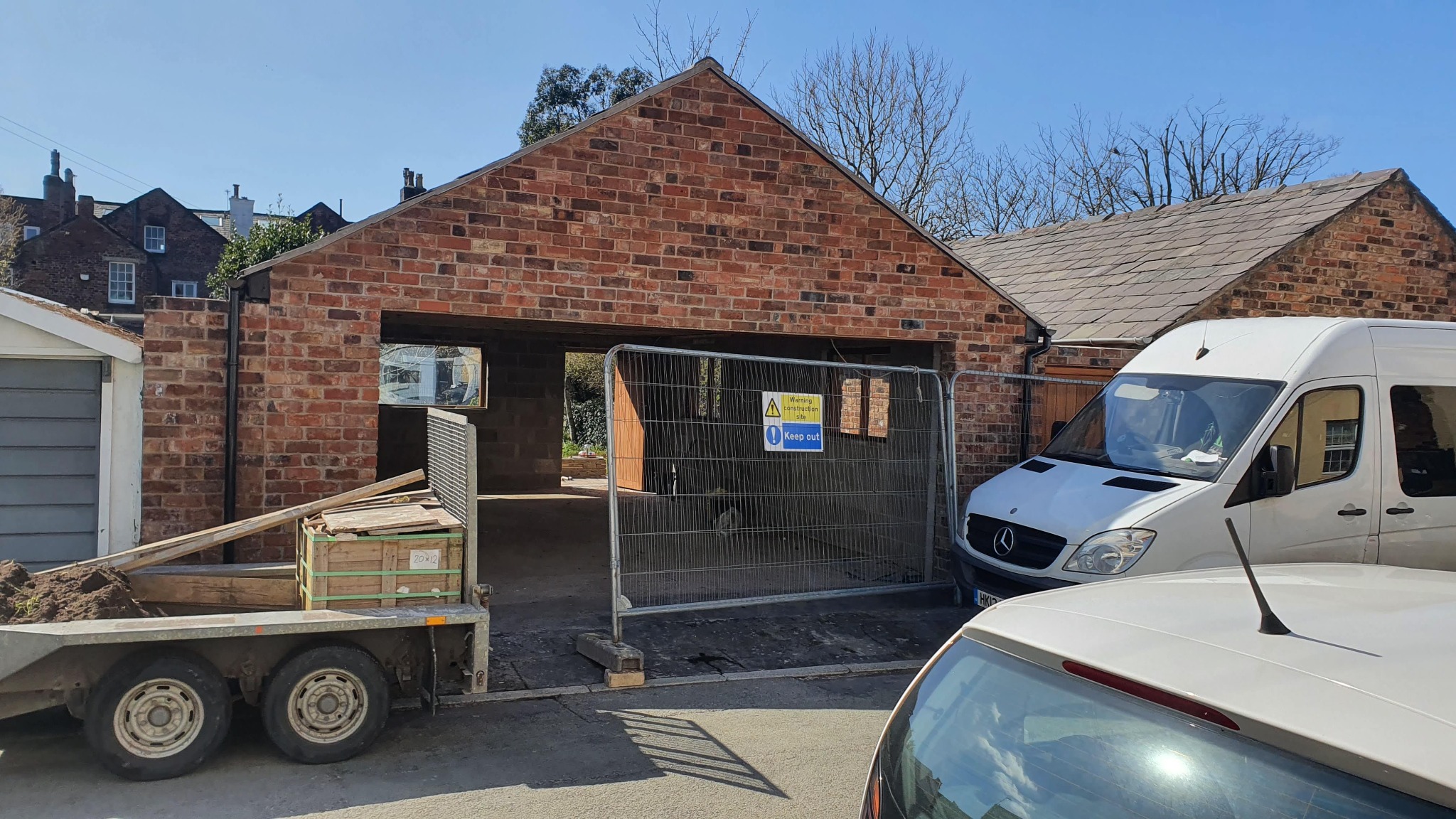 Wznoszona z czerwonej cegły konstrukcja garażu z zabezpieczeniem budowlanym, widoczny przyczepka z materiałami i biały bus na tle zabudowy mieszkalnej.