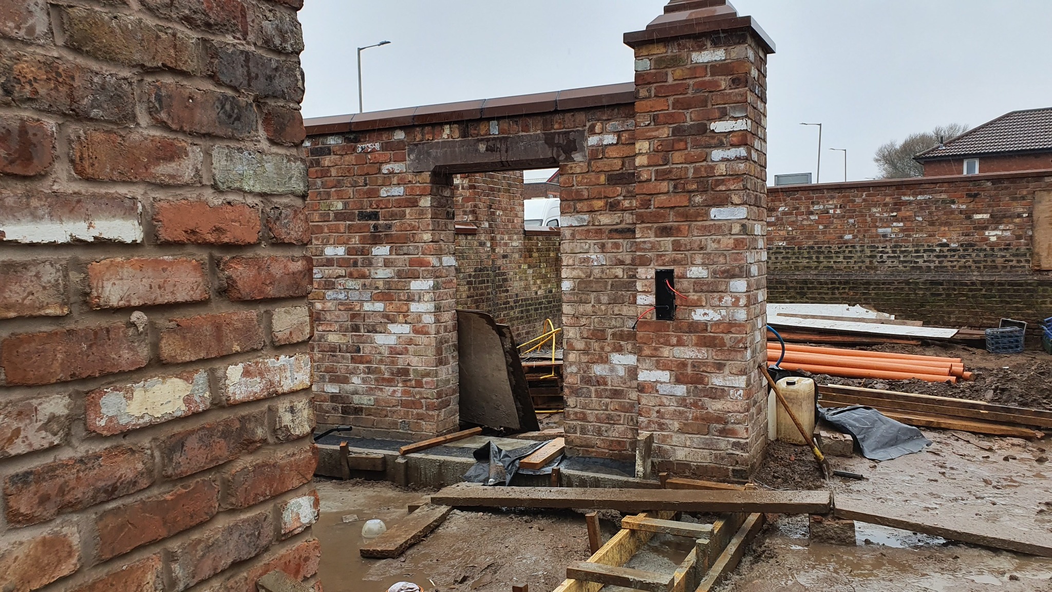 Surowy mur z czerwonej cegły w trakcie wznoszenia, widoczne otwory okienne i drzwiowe, teren budowy z elementami konstrukcyjnymi i narzędziami.