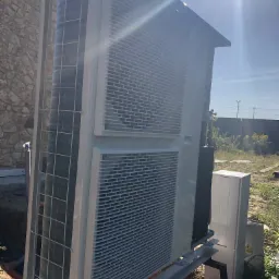 Zewnętrzna jednostka pompy ciepła, zamontowana na metalowej konstrukcji przy budynku z kamienia, widoczna instalacja i skrzynka elektryczna. Słoneczny dzień.