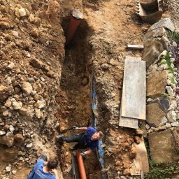 Instalacje Sanitarne WOD-KAN-GAZ-CO Wojciech Olesek - Montaż rur w wykopie ziemnym z użyciem poziomicy. Widoczni dwaj pracownicy w trakcie pracy. W tle fragment koparki i kamienne elementy dekoracyjne.