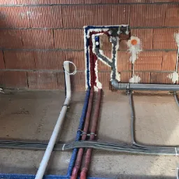 Ściana z pustaków ceramicznych z nową instalacją wodno-kanalizacyjną i grzewczą, rury w kolorach czerwonym i niebieskim, zabezpieczone pianką montażową, widoczne złącza i kształtki.