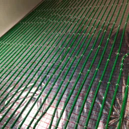 Zielone rury ogrzewania podłogowego ułożone równolegle na srebrnej folii z czerwoną siatką, przymocowane białymi uchwytami, widok pod kątem.