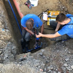 Instalacje Sanitarne WOD-KAN-GAZ-CO Wojciech Olesek - Dwóch instalatorów w niebieskich koszulkach łączy rury w wykopie, używając specjalistycznego narzędzia. Obok leży urządzenie pomiarowe w żółtej obudowie.