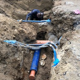 Instalacje Sanitarne WOD-KAN-GAZ-CO Wojciech Olesek - Głęboki wykop w ziemi z ułożonymi rurami instalacyjnymi w kolorze niebieskim i pomarańczowym, pracownik w czarnym kombinezonie pochylony nad instalacją.