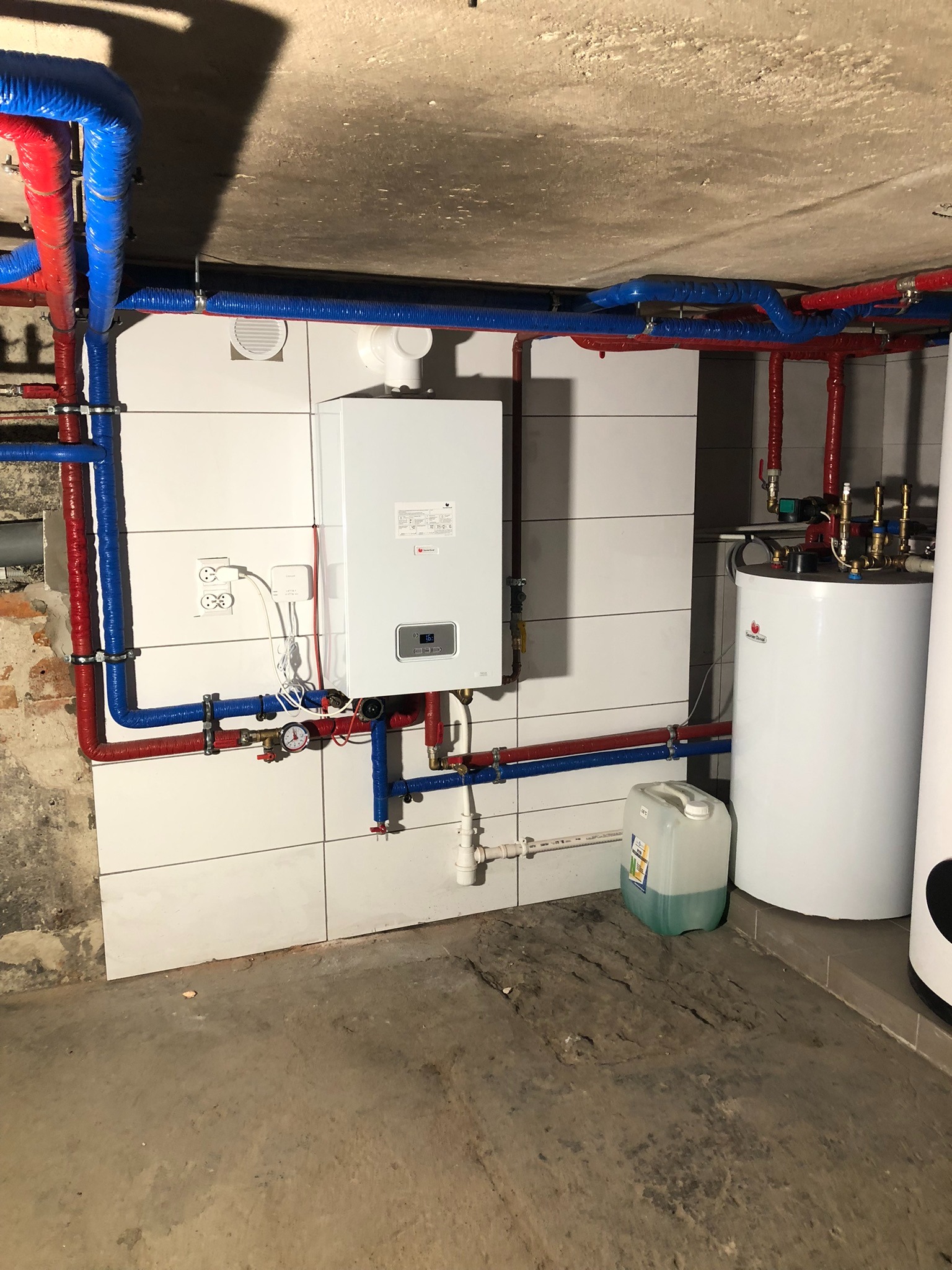 Biały piec gazowy na kafelkowanej ścianie z podłączonymi rurami w kolorze niebieskim i czerwonym, obok zbiornik na wodę i kanister z płynem.