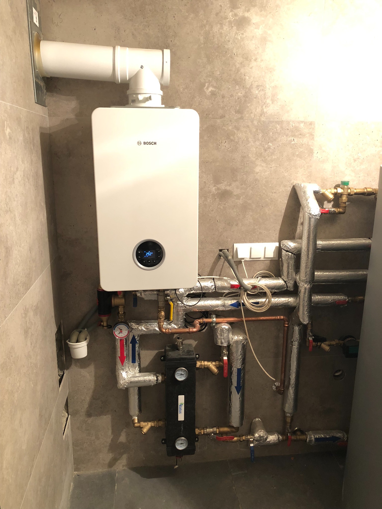 Nowoczesny piec gazowy Bosch z kompletną instalacją hydrauliczną w Myślenicach. Rury zaizolowane, widoczne manometry i zawory. Ściany wykończone płytkami.